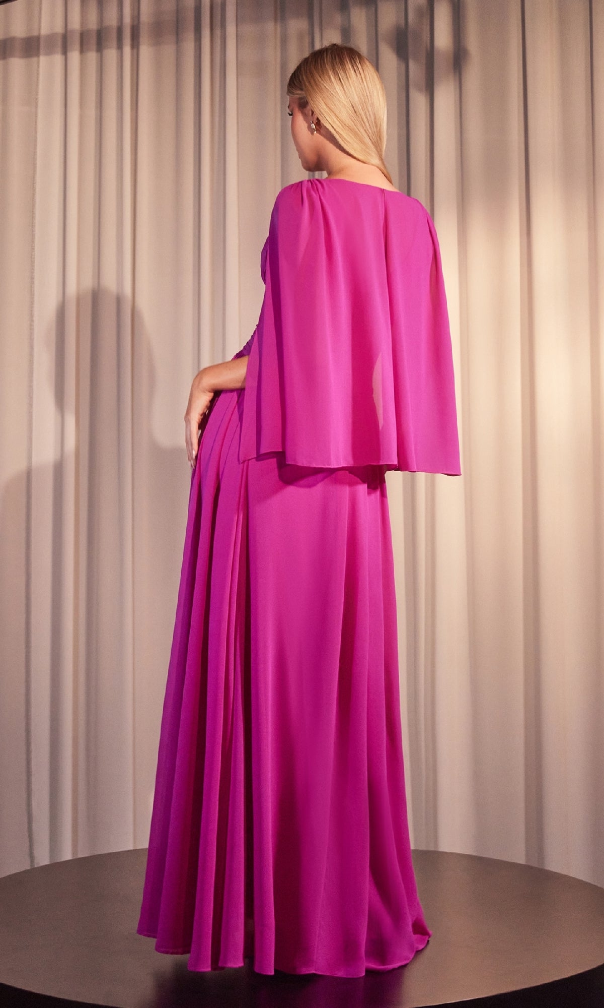 A woman stands on a round platform, facing away from the camera in front of beige curtains, modeling the Ladivine CH028 Ruched Long Chiffon Formal Dress in magenta with a cape.